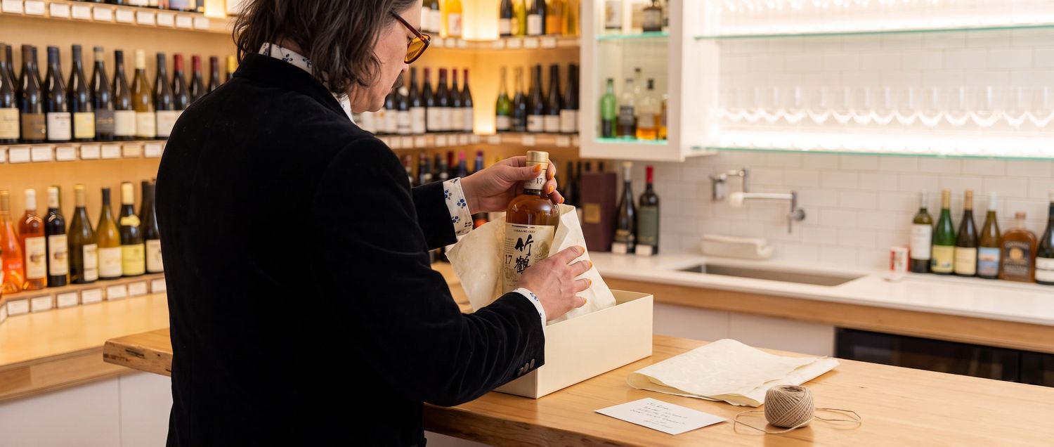 A person in a black jacket lifts a whisky bottle from tissue paper beside a sink and hanging glasses.