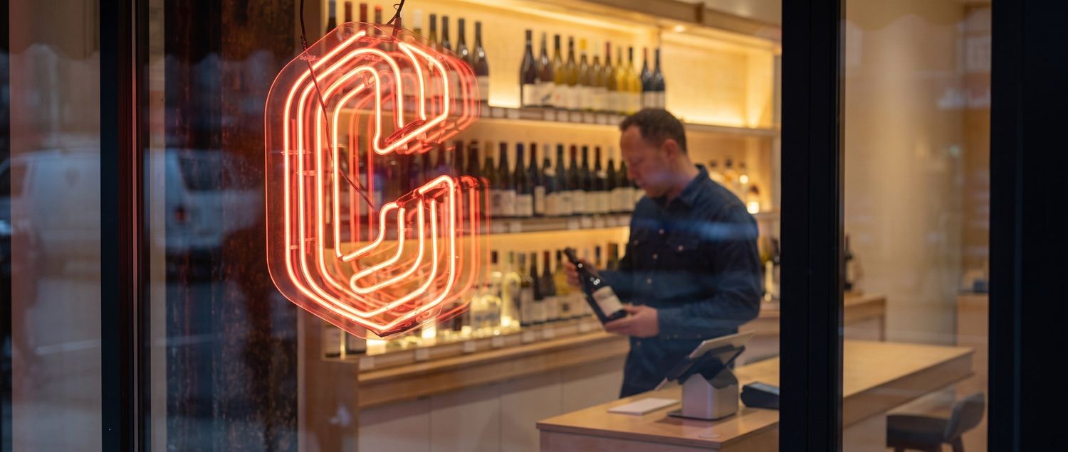 A red neon letter C hangs in a shop window, with a man holding a bottle inside.