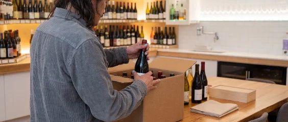A person packs a wine bottle into a plain cardboard box on a pale counter.