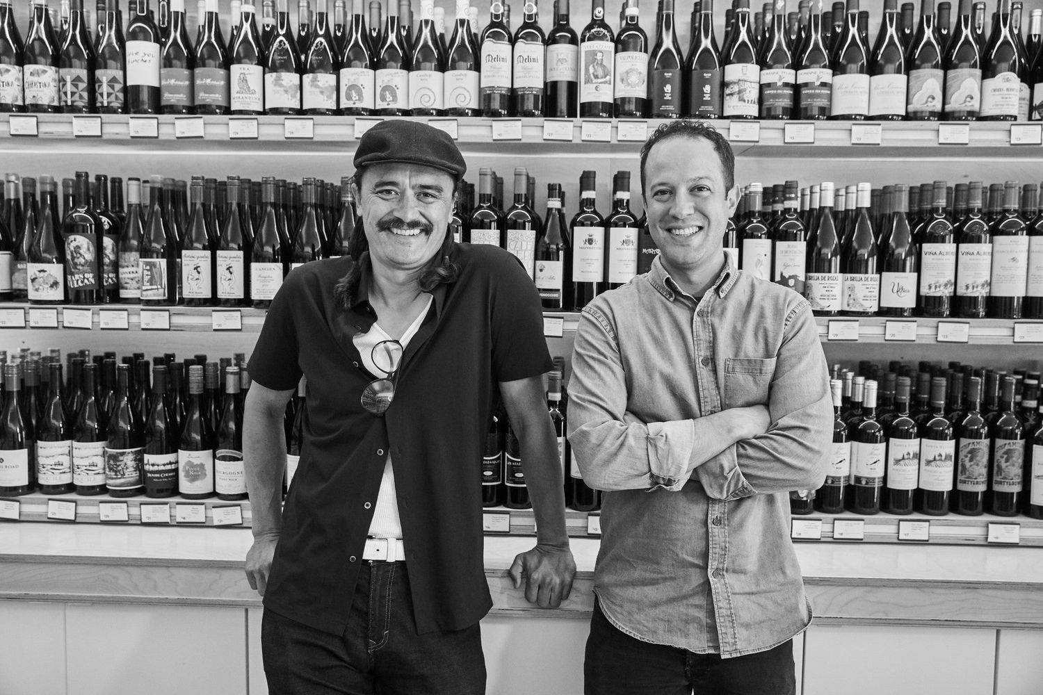 Black and white photograph of two men smiling in front of a dense wall of real wine labels.